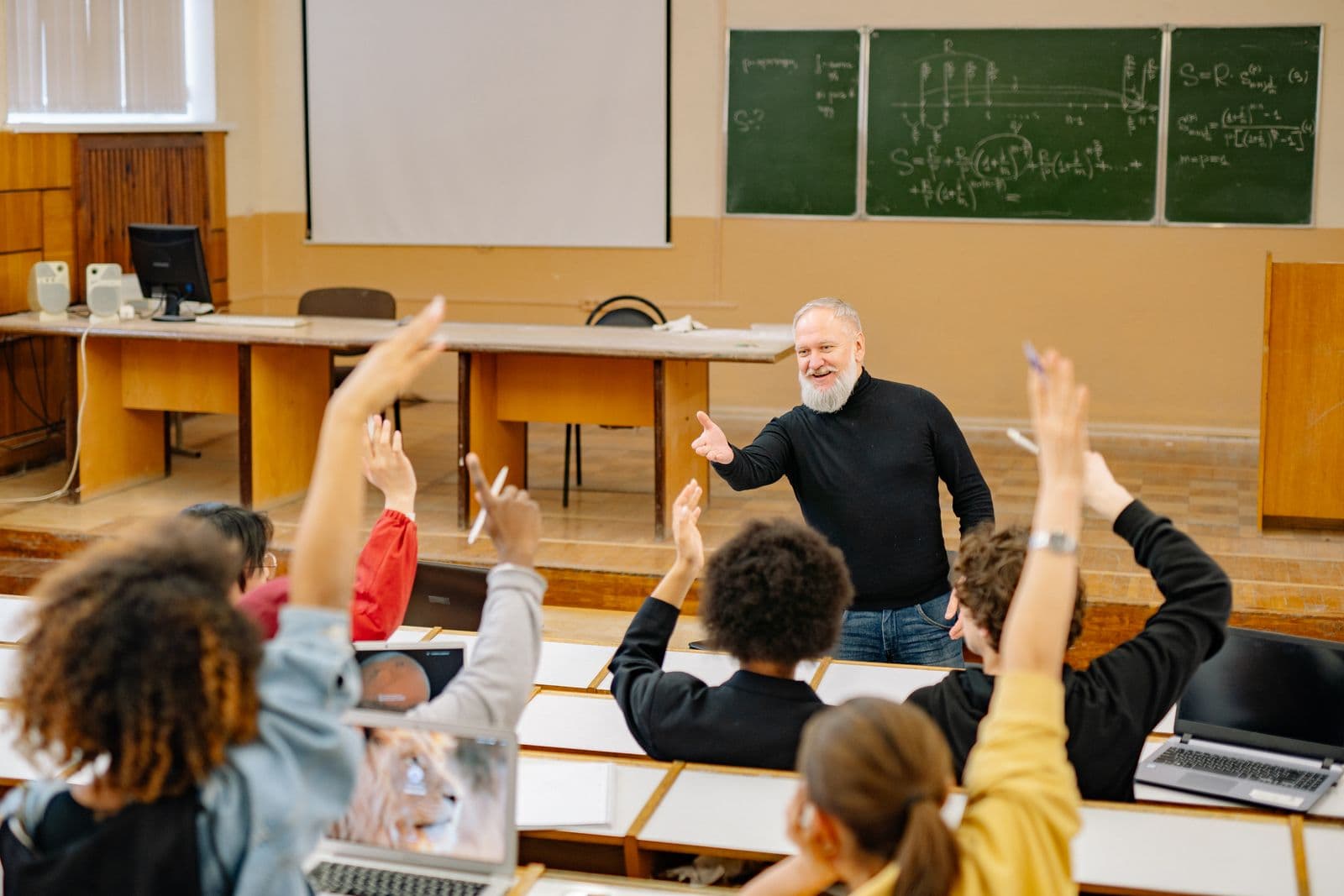 Engaged lecture hall, students raising hands, teacher at blackboard