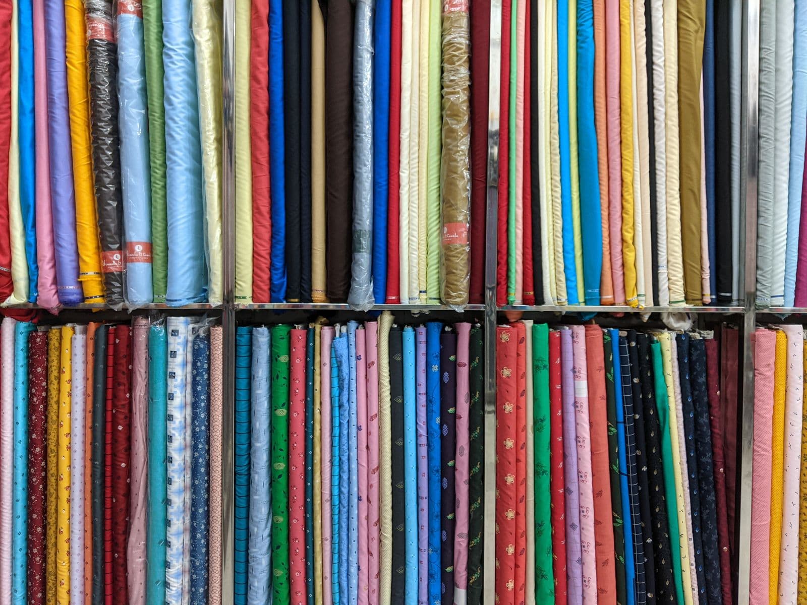 Neat wall of colorful fabric bolts in a cloth shop: solid-colour bolts (uniform fabric) above, printed cottons (fancy/everyday material) below